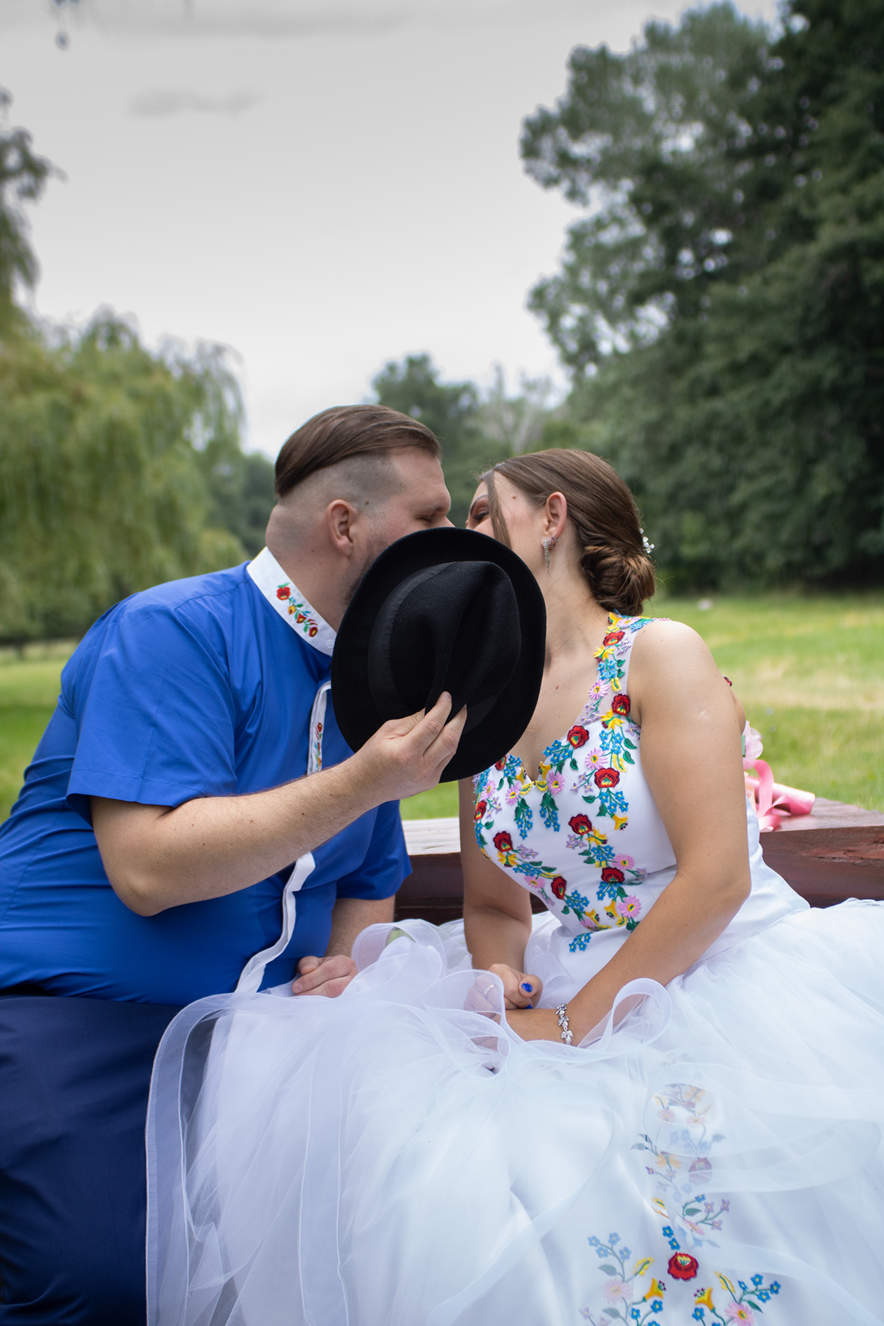 Esküvői fotózás Sástón. A menyasszony és a vőlegény csókját egy fekete kalap takarja el kreatív fotózás közben.